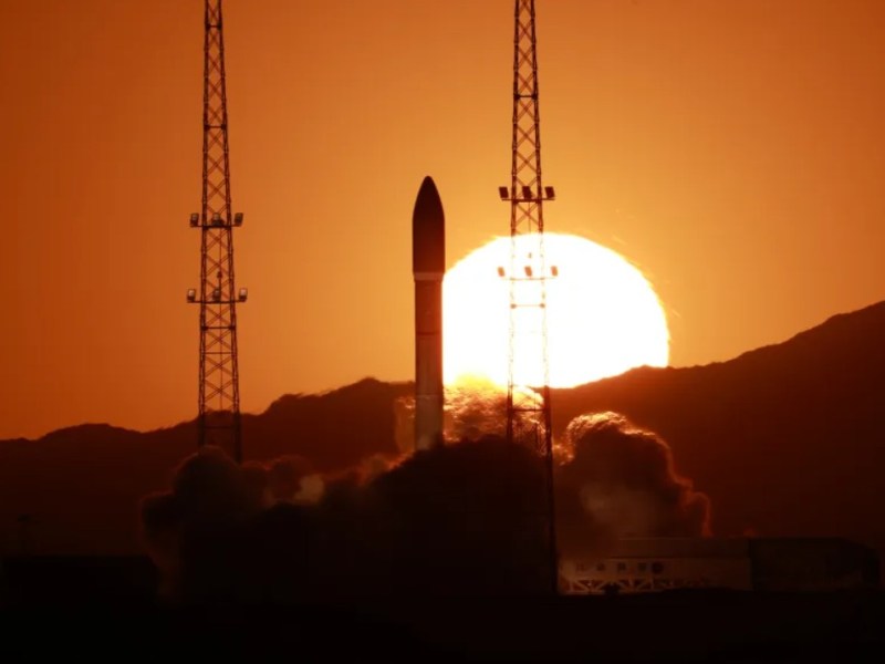 A Ceres-1 solid rocket lifts off from Jiuquan Satellite Launch Center at sunset, framed by two launch towers with the sun glowing behind nearby mountains