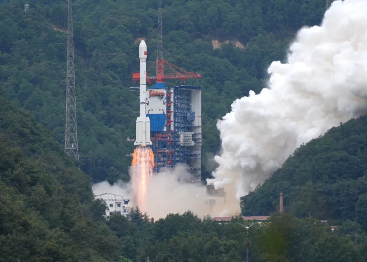 A Long March 3B rocket lifts off from a mountainous launch site in China, engines blazing with bright orange flames and thick white exhaust clouds billowing outward. The vehicle rises beside a tall blue and red service tower, surrounded by dense green forest and hills.