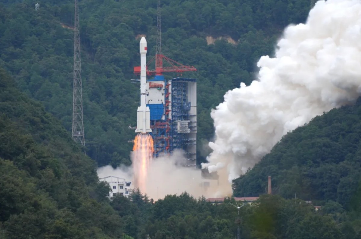 A Long March 3B rocket lifts off from a mountainous launch site in China, engines blazing with bright orange flames and thick white exhaust clouds billowing outward. The vehicle rises beside a tall blue and red service tower, surrounded by dense green forest and hills.