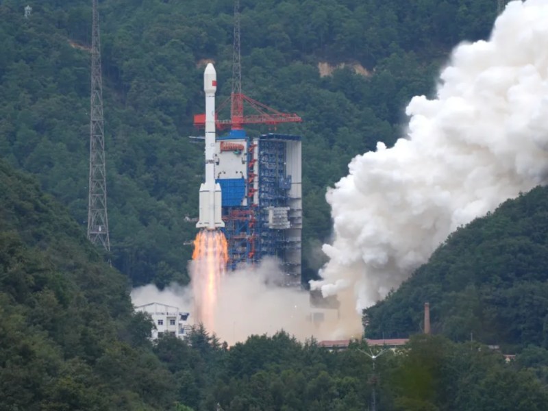A Long March 3B rocket lifts off from a mountainous launch site in China, engines blazing with bright orange flames and thick white exhaust clouds billowing outward. The vehicle rises beside a tall blue and red service tower, surrounded by dense green forest and hills.