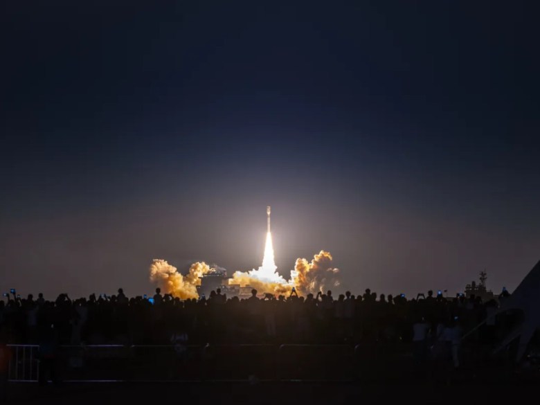 A Jielong-3 rocket lifts off from a sea launch platform at night, its bright exhaust plume illuminating clouds of smoke and silhouetting a crowd of spectators in the foreground.
