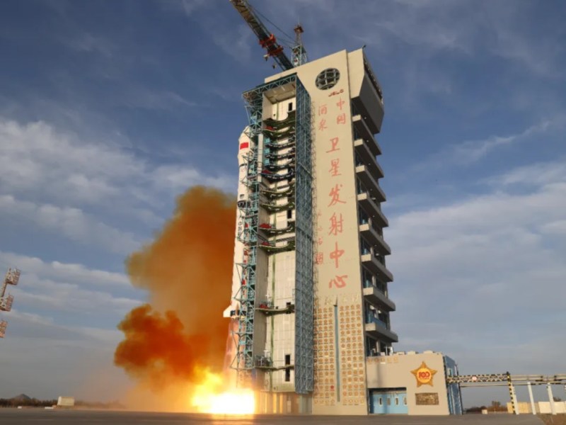 A Long March 2D rocket launches vertically beside a tall, cream-colored service tower at China’s Jiuquan Satellite Launch Center in the Gobi Desert. Thick orange exhaust and flames pour from the rocket’s base, spreading across the gray launch pad. The tower bears large red Chinese characters reading “Jiuquan Satellite Launch Center.” The sky above is pale blue with scattered clouds, and the scene is brightly lit by afternoon sunlight.