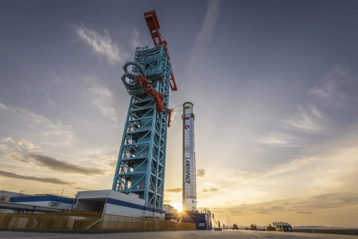 Zhuque-3 first stage stands vertically on the launch pad at Jiuquan Satellite Launch Center in northwest China, secured to a large blue service tower, with the sun setting behind it. The stainless steel rocket displays the Landspace logo prominently.
